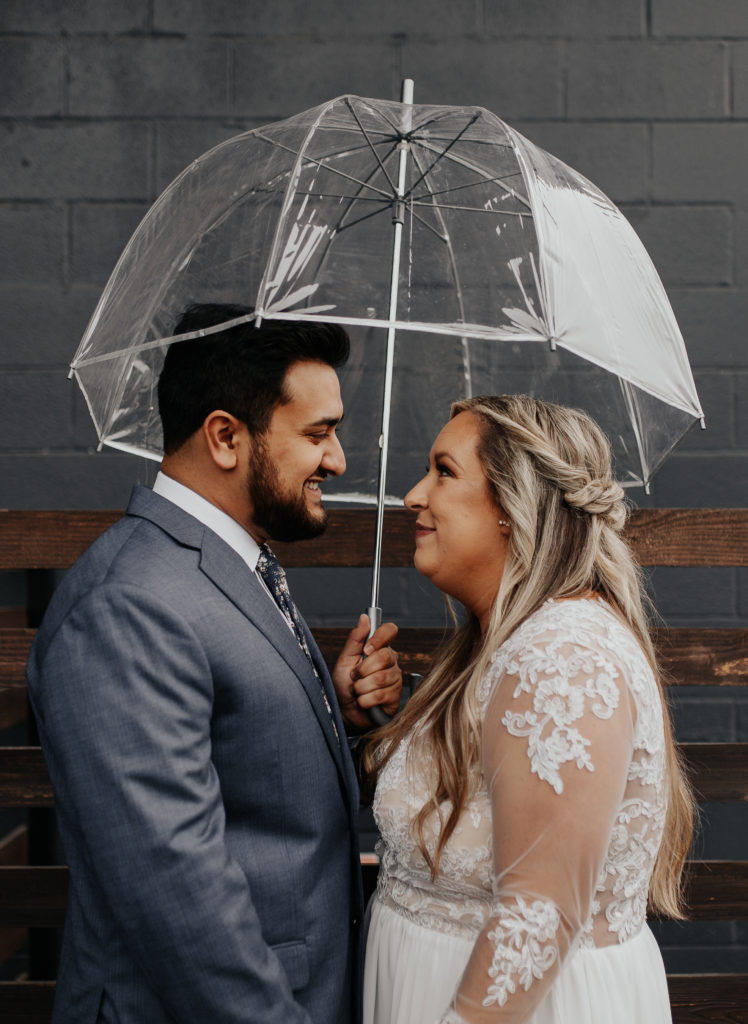 Bride and groom under clear umbrella in front of brick wall